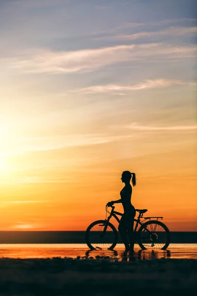 Sylwetka dziewczynki sportowy w garniturze, który siedzi na rowerze w wodzie o zachodzie słońca na ciepły, letni dzień. Koncepcja fitness. Tło nieba — Zdjęcie stockowe