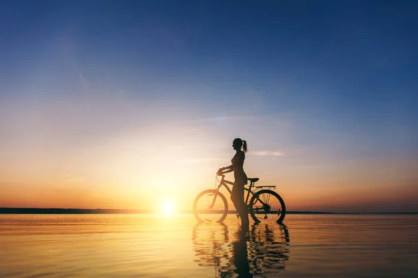 Silhouette of a sporty girl in a suit that sits on a bicycle in the water at sunset on a warm summer day. Fitness concept. Sky background — Stock Photo, Image