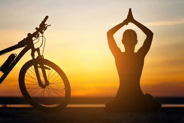 Sports woman doing yoga at sunrise on the sea beach against the background of orange sky and bicycle. Fitness concept.