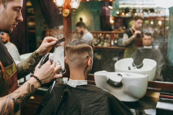 Um homem a cortar o cabelo na barbearia. Cabeleireiro masculino em tatuagens servindo cliente . — Fotografia de Stock