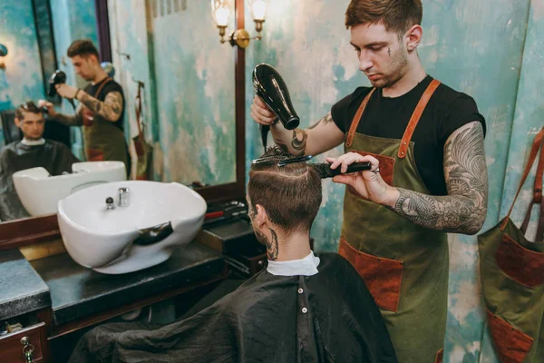 Mann bekommt trendigen Haarschnitt beim Friseur. männlicher Friseur mit Tätowierungen im Dienste des Kunden, Föhnen der Haare mit einem Haartrockner — Stockfoto