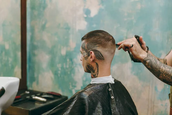 Close up shot of man getting trendy haircut at barber shop. Male hairstylist in tattoos serving client. — Stock Photo, Image