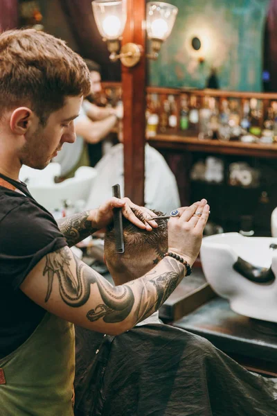 Um homem a cortar o cabelo na barbearia. Cabeleireiro masculino em tatuagens servindo cliente . — Fotografia de Stock