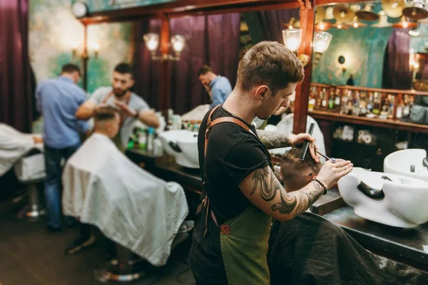 Um homem a cortar o cabelo na barbearia. Cabeleireiro masculino em tatuagens servindo cliente . — Fotografia de Stock