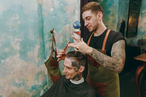 Um homem a cortar o cabelo na barbearia. Cabeleireiro masculino em tatuagens servindo cliente . — Fotografia de Stock