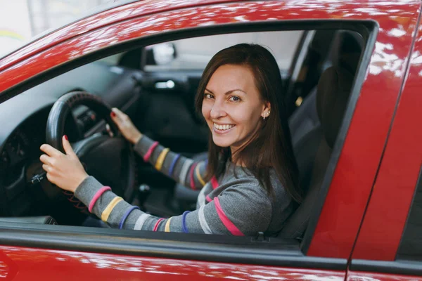 Beautiful young happy smiling European brown-haired woman with healthy clean skin dressed in a striped t-shirt sits in her red car with black interior. Traveling and driving concept. Royalty Free Stock Photos