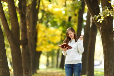 Güzel bir mutlu gülümseyen kahverengi saçlı kadın beyaz kazak daimi sıcak bir günde sonbahar Şehir Parkı içinde kırmızı bir kitapla. Altın yaprak. Okuma kavramı.