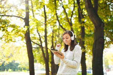 Güzel bir mutlu neşeli kahverengi saçlı Bayan Beyaz kazaklı bir tablet sonbahar Park'ta sıcak bir gün beyaz kulaklık müzik dinleme. Şehirde sonbahar.