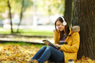 Bir kadında sarı ceket ve kot bardak kahve veya çay ile oturup ve sıcak bir günde bir tablet onun elinde ve sonbahar Şehir Parkı kulaklık ile bir ağacın altında müzik dinlemek. Altın yaprak.