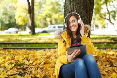 Kahverengi saçlı gülümseyen kadın sarı ceket ve kot oturan ve sıcak bir günde bir tablet onun elinde ve sonbahar Şehir Parkı kulaklık ile bir ağacın altında müzik dinlemek. Altın yaprak.
