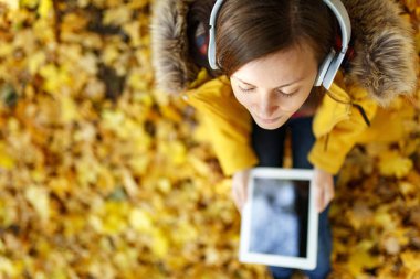 Sarı ceket ve oturma ve sıcak bir günde bir tablet onun elinde ve sonbahar Şehir Parkı kulaklık ile bir ağacın altında müzik dinlemek kot kahverengi saçlı bir kadın. Altın yaprak. Üstten Görünüm