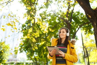 Güzel bir mutlu neşeli kahverengi saçlı kadın sarı ceket ve çizgili longsleeve bir tablet ile sıcak bir günde ellerini ve beyaz kulaklık sonbahar Şehir Parkı içinde sevinir. Altın yaprak.