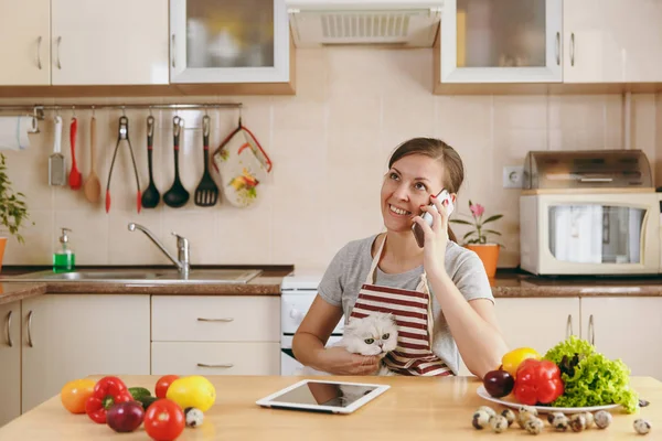 Una giovane bella donna con gatto bianco persiano che parla al cellulare in cucina con tablet sul tavolo. Insalata di verdure. Il concetto di dieta. Uno stile di vita sano. Cucinare a casa. Preparare il cibo . — Foto Stock