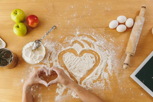 Hand dras hjärtat i mjöl på köksbordet och andra ingredienser och tablett. Ovanifrån. — Stockfoto