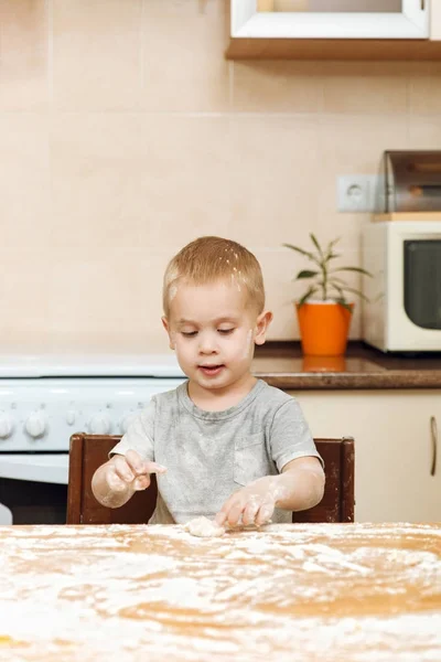 Маленький мальчик помогает маме готовить рождественское имбирное печенье на светлой кухне. Счастливый светловолосый ребенок в серой футболке 2-3 года за столом с тестом и мукой в выходные утром дома . — стоковое фото