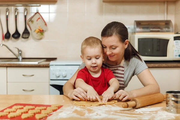 Little kid boy helps mother to cook Christmas ginger biscuit in light kitchen. Happy family mom 30-35 years and child 2-3 roll out dough and cut out cookies at home. Relationship and love concept