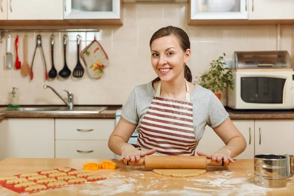 Une belle jeune femme heureuse 30-35 ans dans tablier assis à une table avec de la pâte et du rouleau à pâtisserie et va préparer un gâteau au gingembre de Noël dans la cuisine légère. Je cuisine à la maison. Préparer les aliments . — Photo