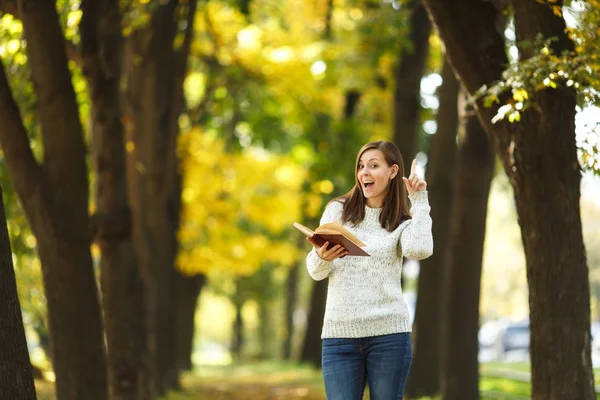 A beautiful happy smiling brown-haired woman in white sweater standing with a red book in fall city park on a warm day. Autumn golden leaves. Reading concept.