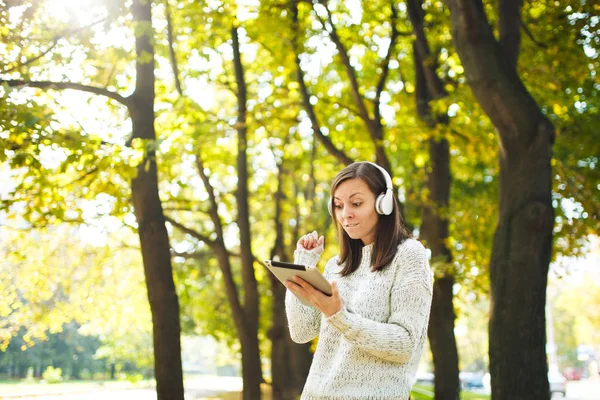 Μια όμορφη ευτυχισμένη χαρούμενη καστανά μαλλιά γυναίκα στο λευκό πουλόβερ με ένα δισκίο ακούγοντας μουσική στα λευκά ακουστικά σε πτώση πάρκο σε μια ζεστή μέρα. Φθινόπωρο στην πόλη. — Φωτογραφία Αρχείου