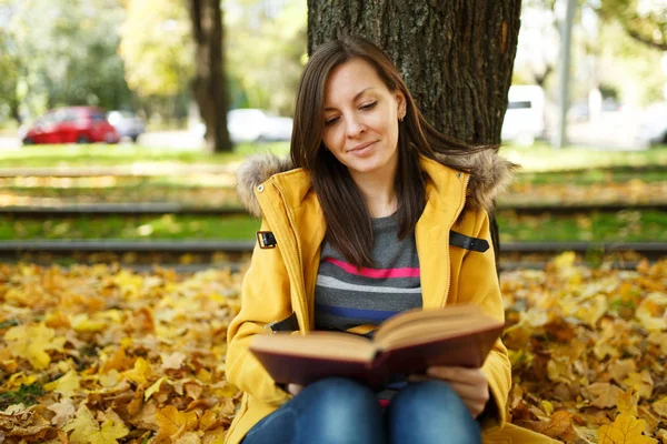 Piękny szczęśliwy uśmiechający się Brunet Kobieta w żółtym fartuchu i dżinsy, siedząc pod drzewem klonowym czerwony książkę w Upadek miasta park na ciepły dzień. Autumn golden odchodzi. Koncepcja czytania — Zdjęcie stockowe