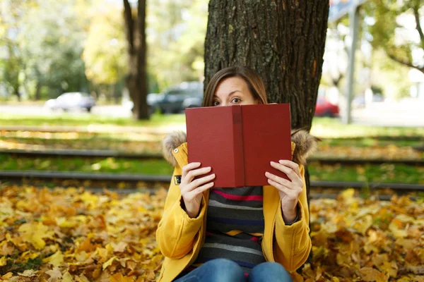 A beautiful happy smiling brown-haired woman in yellow coat and jeans sitting under the maple tree and hiding behind red book in fall city park on a warm day. Autumn golden leaves. Reading concept