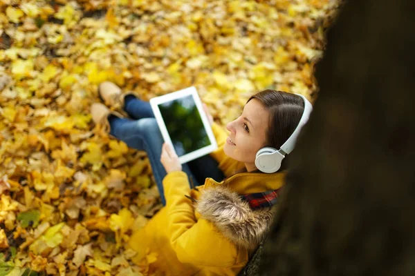 Καστανά μαλλιά γυναίκα σε κίτρινο παλτό και τζιν κάθεται και ακούγοντας μουσική κάτω από ένα δέντρο με ένα tablet στα χέρια της και ακουστικά στο πάρκο πόλης πτώση σε μια ζεστή μέρα. Χρυσή φθινόπωρο φύλλα. Το Top view — Φωτογραφία Αρχείου