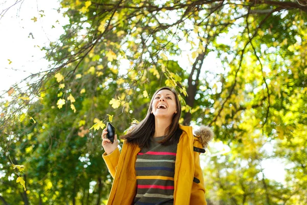 A beautiful happy cheerful brown-haired woman in a yellow coat and striped longsleeve talking on mobile phone in fall city park on a warm day. Autumn golden leaves.