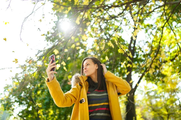 A beautiful happy cheerful brown-haired woman in a yellow coat and striped longsleeve doing selfie on mobile phone in fall city park on a warm day. Autumn golden leaves.