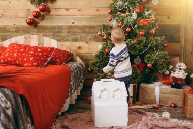 Mutlu sevimli küçük erkek bebek kazak ve ahşap Oda evde oyuncaklar ile Noel ağacı süsleme kot pantolon giymiş. Çocuk iyi bir ruh hali ile. Yeni yıl. Yaşam tarzı, Aile ve tatil 2018 kavramı