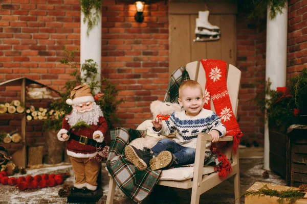 Juguetón niño lindo feliz vestido con suéter y vaqueros sentados en la silla con Santa en la habitación decorada de Año Nuevo en casa. Navidad buen humor. Estilo de vida, familia y vacaciones 2018 concepto — Foto de Stock