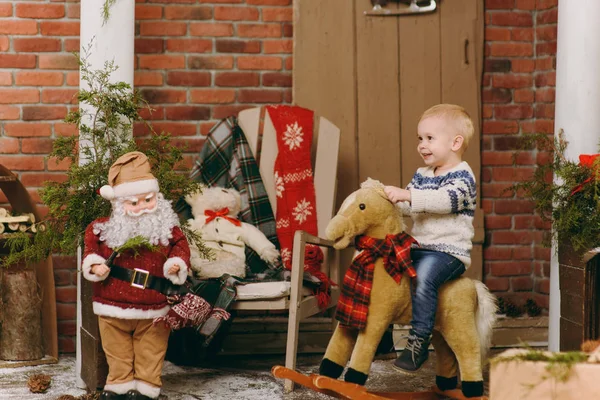 Juguetón niño lindo feliz vestido con suéter y vaqueros sentados en el caballo mecedora en la habitación decorada de Año Nuevo con Santa en casa. Navidad buen humor. Estilo de vida, familia y vacaciones 2018 concepto — Foto de Stock