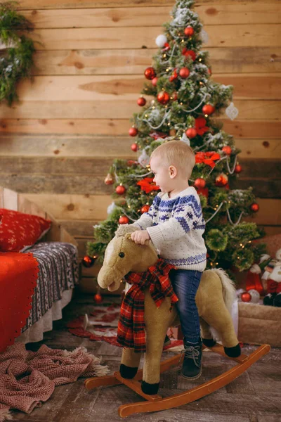Feliz lindo niño en el caballo mecedora en el árbol de Navidad con un juguete, vestido con suéter y vaqueros en la habitación decorada de Año Nuevo en casa. Buen humor para niños. Estilo de vida, familia y vacaciones 2018 concepto — Foto de Stock