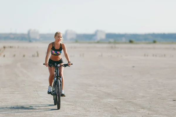 Uma mulher loira esportiva em um terno colorido anda de bicicleta em uma área deserta em um dia ensolarado de verão. Conceito de aptidão. Céu azul fundo — Fotografia de Stock