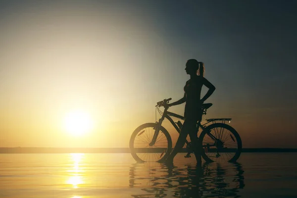 Silna Kobieta blonde w kolorowy komplet stoi w pobliżu rowerów w wodzie o zachodzie słońca na ciepły, letni dzień. Koncepcja fitness. Tło nieba — Zdjęcie stockowe