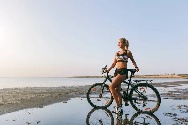 Uma mulher loira forte em um terno colorido fica perto de uma bicicleta em uma área deserta perto da água. Conceito de aptidão. Céu azul fundo — Fotografia de Stock