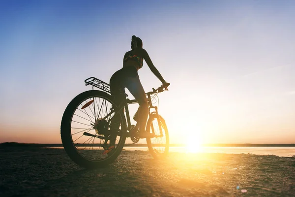 A strong blonde woman in a multicolored suit sits on a bicycle in a desert area near the water and looks at the sun. Fitness concept. Back view — Stock Photo, Image
