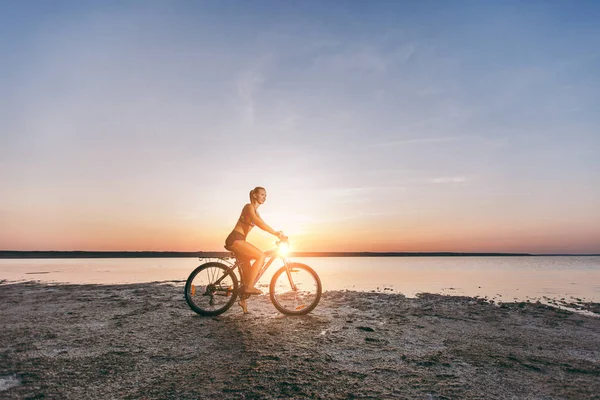 Sportowy blond kobieta w kolorowy komplet przejażdżki rowerowe w okolicy pustyni w pobliżu wody w słoneczny letni dzień. Koncepcja fitness. Na tle niebieskiego nieba — Zdjęcie stockowe