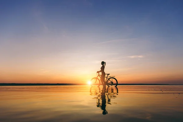 Silhouette of a sporty girl in a suit that sits on a bicycle in the water at sunset on a warm summer day. Fitness concept. Sky background — Stock Photo, Image