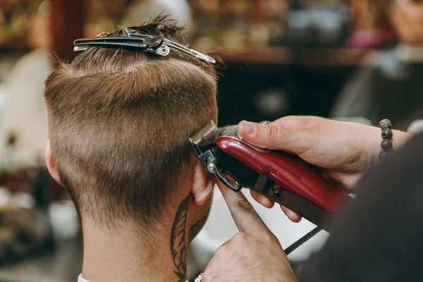 Tiro de perto de um homem a cortar o cabelo na barbearia. Cabeleireiro masculino em tatuagens servindo cliente . — Fotografia de Stock