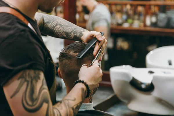 Tiro de perto de um homem a cortar o cabelo na barbearia. Cabeleireiro masculino em tatuagens servindo cliente . — Fotografia de Stock