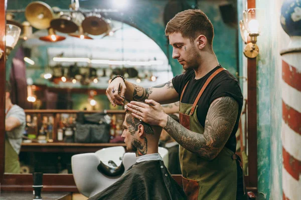 A man getting trendy haircut at barber shop. Male hairstylist in tattoos serving client. — Stock Photo, Image