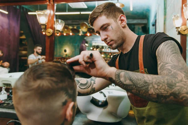 Tiro de perto de um homem a cortar o cabelo na barbearia. Cabeleireiro masculino em tatuagens servindo cliente . — Fotografia de Stock