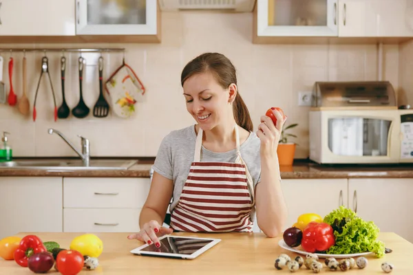 Una giovane donna seduta accanto al tavolo e alla ricerca di una ricetta nella tavoletta in cucina. Insalata di verdure. Il concetto di dieta. Uno stile di vita sano. Cucinare a casa. Preparare il cibo . — Foto Stock