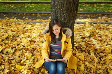 Güzel bir mutlu gülümseyen kahverengi saçlı kadın sarı ceket ve kot sıcak bir günde sonbahar Şehir Parkı kırmızı bir kitap ile akçaağaç ağacın altında oturuyor. Altın yaprak. Okuma kavramı
