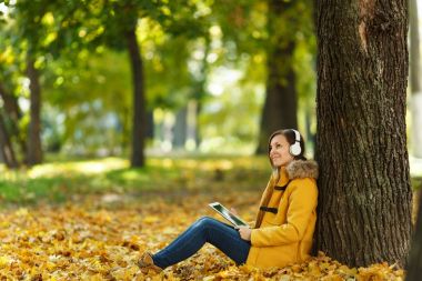 Kahverengi saçlı gülümseyen kadın sarı ceket ve kot oturan ve sıcak bir günde bir tablet onun elinde ve sonbahar Şehir Parkı kulaklık ile bir ağacın altında müzik dinlemek. Altın yaprak.