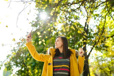 Güzel bir mutlu neşeli kahverengi saçlı kadın sarı ceket ve çizgili longsleeve sonbahar Şehir Parkı cep telefonunda selfie sıcak bir günde yapıyor. Altın yaprak.
