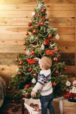 Mutlu sevimli küçük erkek bebek kazak ve ahşap Oda evde oyuncaklar ile Noel ağacı süsleme kot pantolon giymiş. Çocuk iyi bir ruh hali ile. Yeni yıl. Yaşam tarzı, Aile ve tatil 2018 kavramı