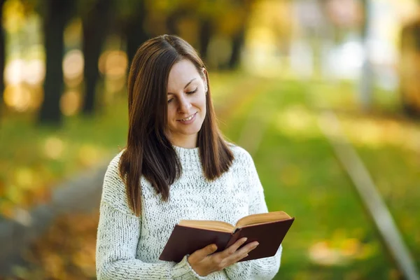 Uma bela mulher sorridente feliz de cabelos castanhos em camisola branca de pé com um livro vermelho nas trilhas de bonde no parque da cidade de outono em um dia quente. Folhas douradas de outono. Conceito de leitura . — Fotografia de Stock