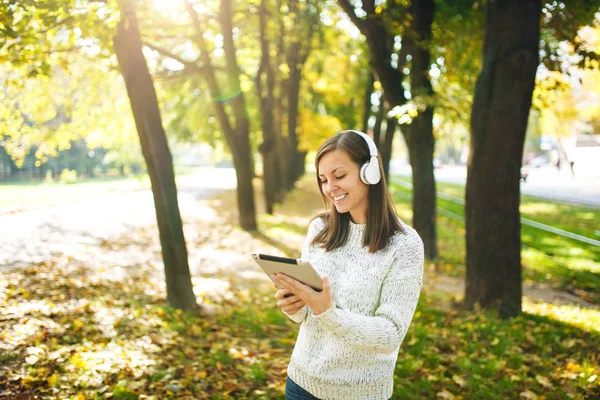 Güzel bir mutlu neşeli kahverengi saçlı Bayan Beyaz kazaklı bir tablet sonbahar Park'ta sıcak bir gün beyaz kulaklık müzik dinleme. Şehirde sonbahar.