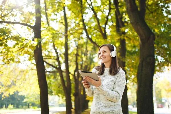 A beautiful happy cheerful brown-haired woman in white sweater with a tablet listening music in the white headphones in fall park on a warm day. Autumn in the city.
